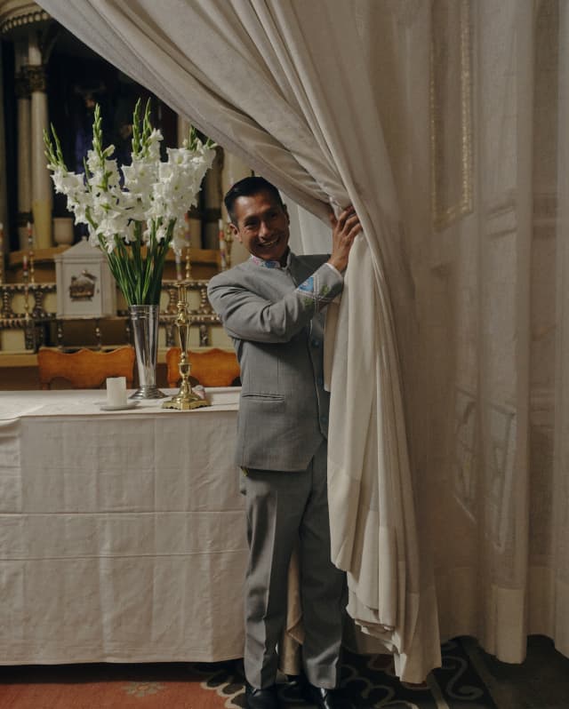 A man in a grey suit pulls drapes aside to reveal a clothed table with a vase of gladioli in the Palacio Nazarenas chapel.