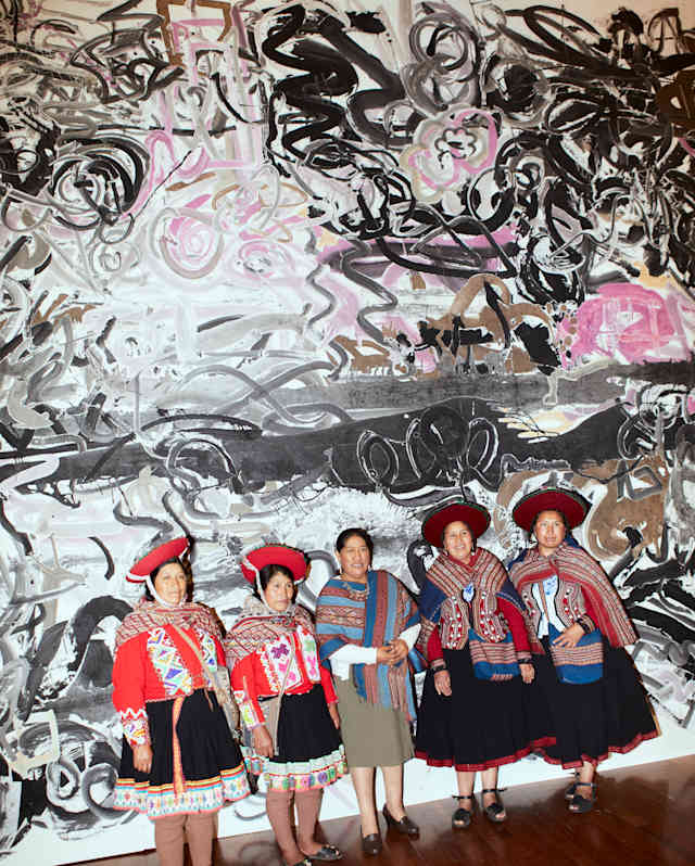 Five people wearing colourful traditional Andean clothing stand in front of a large abstract black, white, and pink painting on a gallery wall with a wooden floor.