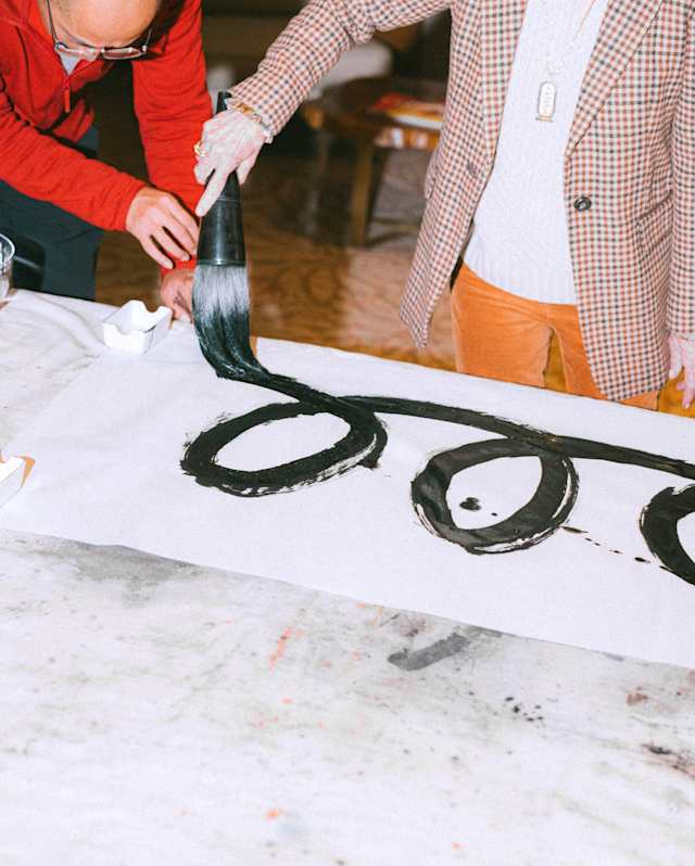 Two people are collaboratively painting large, black, circular shapes on a white canvas spread across a table. Their faces are not visible, and one person holds a brush whilst the other assists.