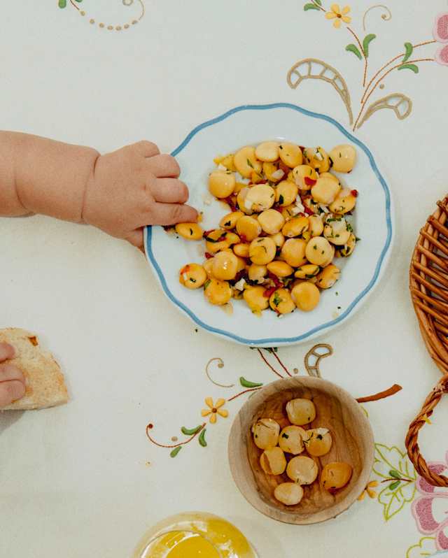 A small child’s hand reaches for a plate of chickpea salad on a table with a floral tablecloth. Nearby are a bowl with more chickpeas, a lemon drink, and a piece of bread.