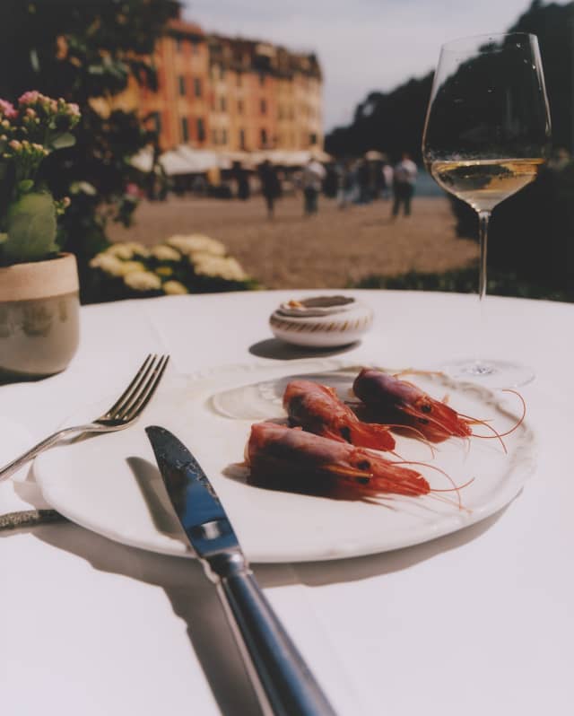 A knife and fork rest against a plate with three Santa Margherita prawns at a table with white wine set back from the marina.