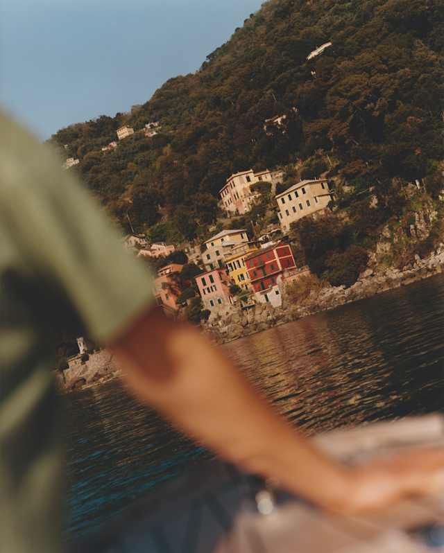Angled image of Portofino's red, pink and yellow marina-side buildings, seen over the arm of a man on a boat in soft-focus.