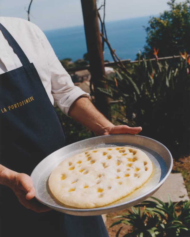 A chef in a dark apron presents a flat, doughy Focaccia Genovese bread on a silver tray ready for baking, seen close-up.