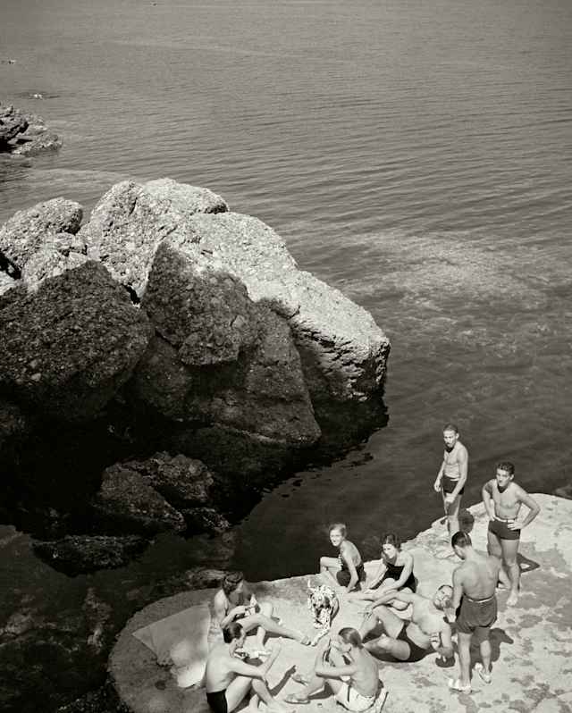 1936 monochrome photo of a group of men and women in swimming suits basking on a rock with a Dalamation, by Herbert List.