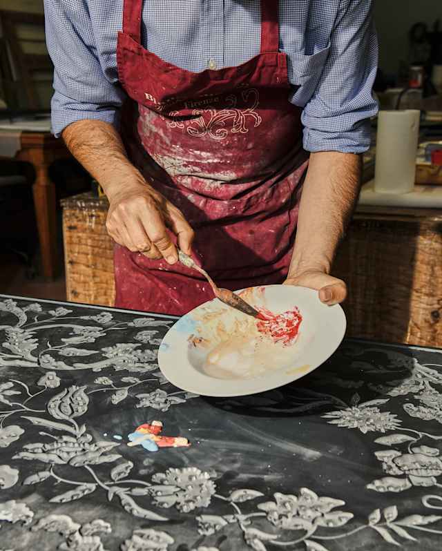 Bianco Bianchi, seen from the neck down in a red apron, prepares a Scagliola plaster paste to add inlay to an etched artwork.