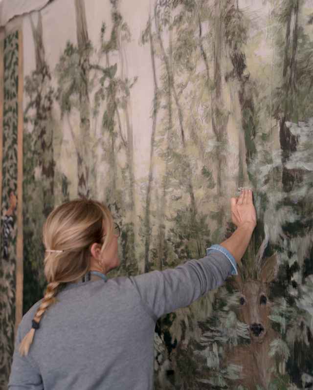 Elena Carozzi, with blond hair in a plait, rests her hand on a large hanging paper with botanical motifs, seen from the side.