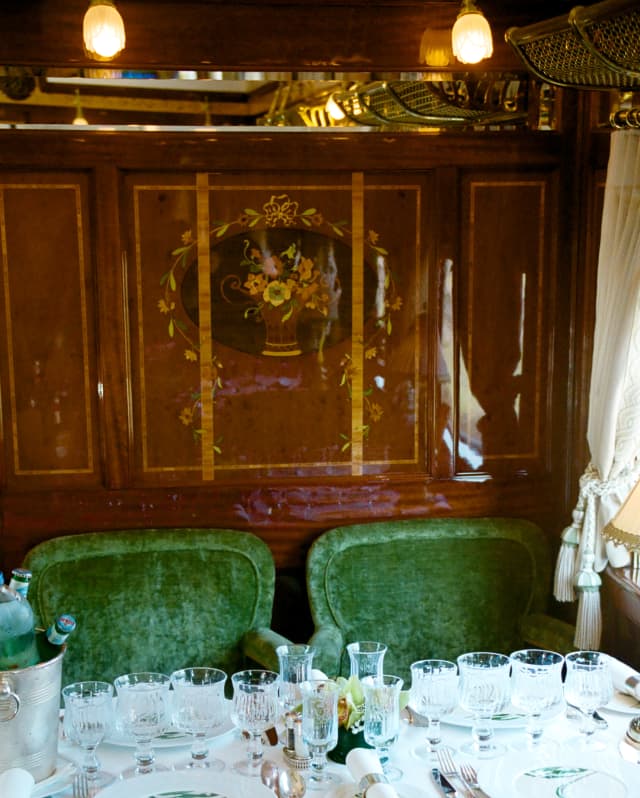 Two plush green chairs sit beneath a floral marquetry panel, seen over a table filled with glasses in the Etoile du Nord car.