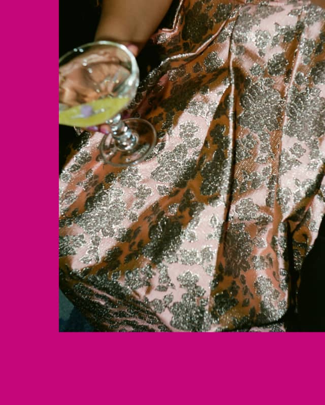 A woman holds a drink on the lap of her rose silken skirt with floral metallic brocade, pictured close-up on pink background.