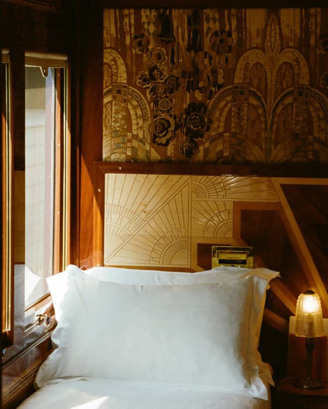 A single bed in crisp white linen with a propped pillow settles between a cabin window and a side table with a glowing lamp.
