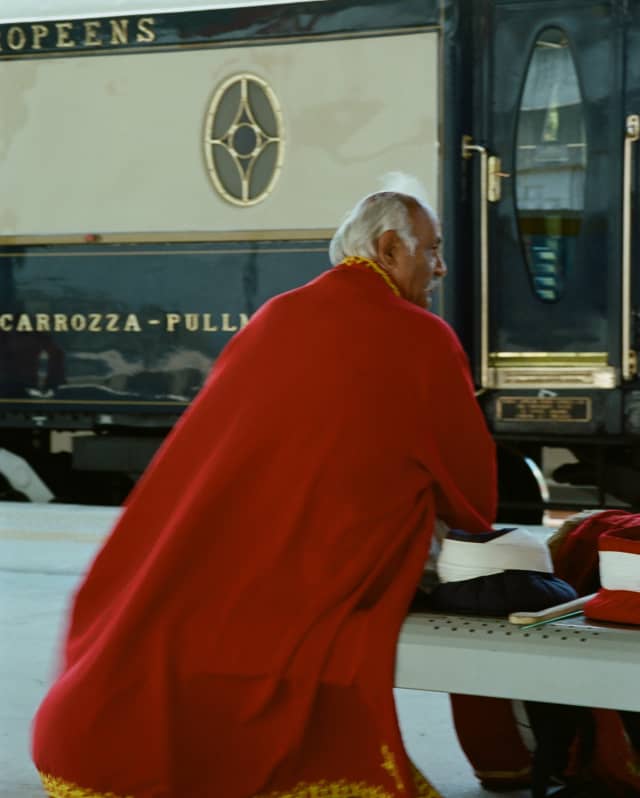 A grey-haired man sits on a platform bench in front of the train, in a long red cloak with gold piping, seen from behind.