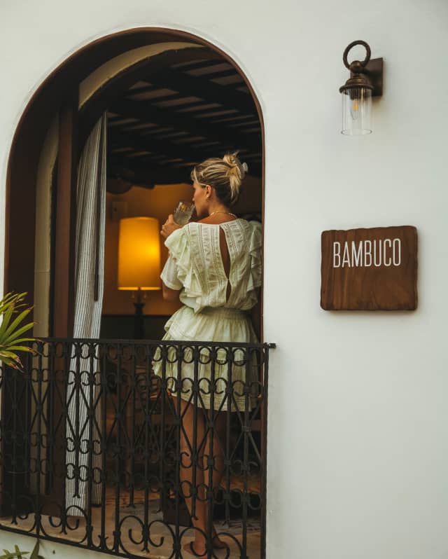 Bambuco's wood sign adorns the exterior wall by an open arched window where a blond woman in a white dress sips from a glass.