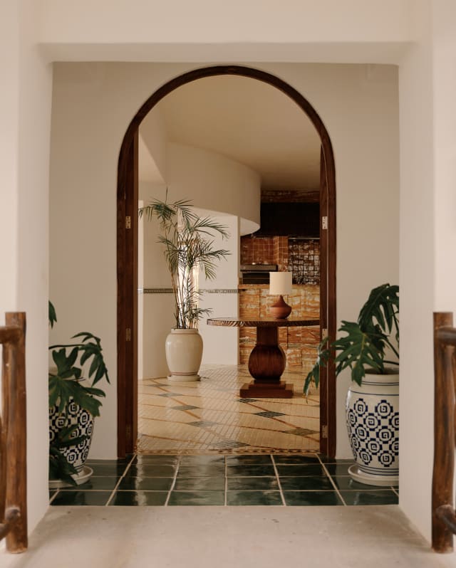 Potted monstera plants guard the archway entrance to Woodend with a wooden door frame and glazed green-tile threshold.
