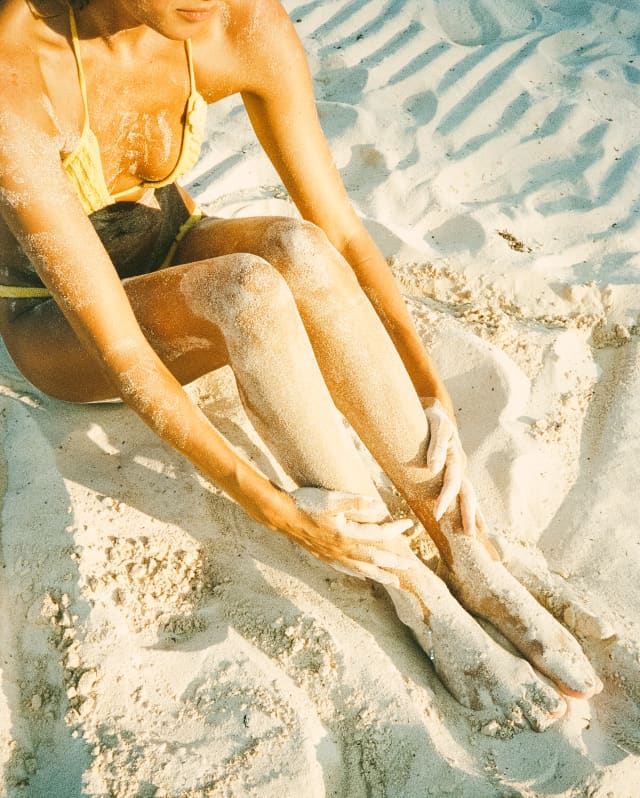 Close-up of a woman in a yellow bikini as she sits on the beach with her legs covered in sand, seen from above.
