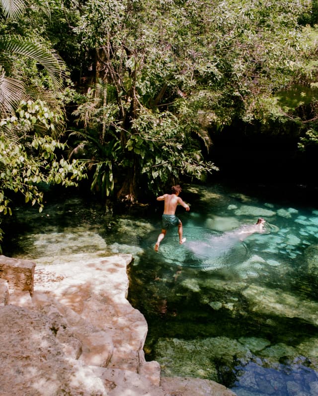 A man leaps of the pink rock edge of an open cenote where a friend already swims in the incredible turquoise waters.