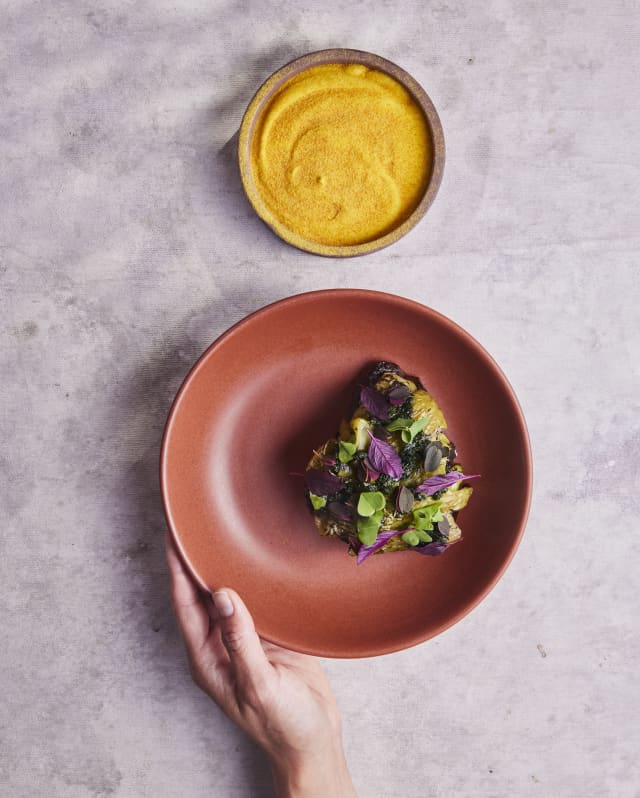 A hand holds a reddish-brown plate with a small salad of greens and purple leaves, next to a small bowl of yellow sauce or dip, set on a light grey surface.
