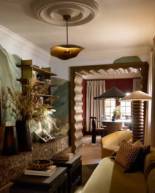 A cosy living room with a green sofa, cushions, coffee tables, built-in shelves with vases and books, sculpted wall detailing, and warm ambient lighting from ceiling and table lamps. A window lets in natural light in the background.