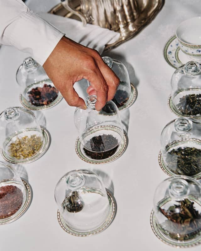 The hand of a tea sommelier selects one of nine varieties of tea leaves, displayed on small plates beneath glass cloches.