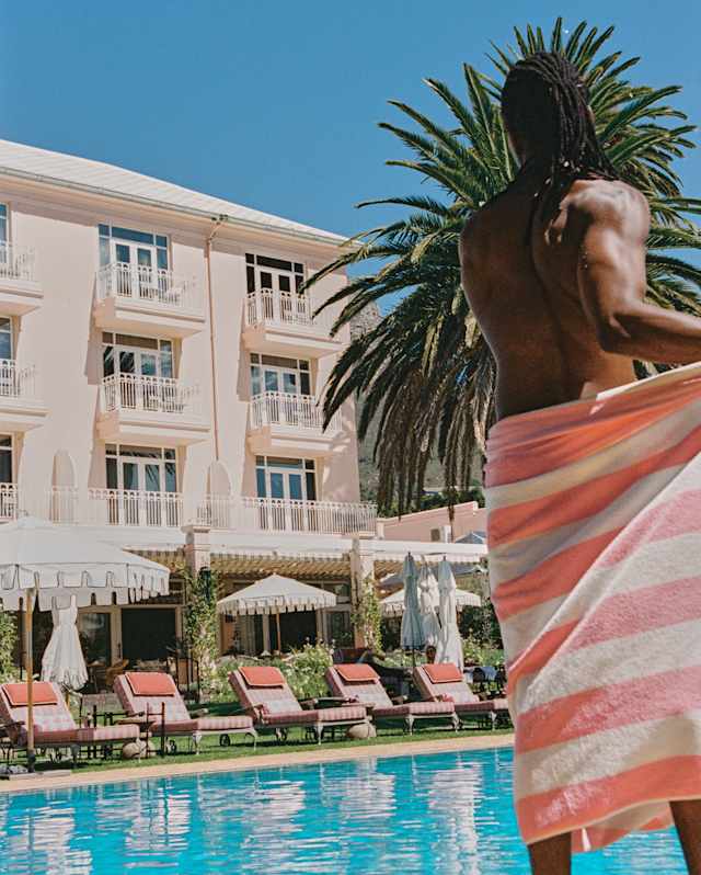 A person with long hair stands by a pool wrapped in a pink and white striped towel. Behind them are sun loungers, parasols, palm trees, and a light-coloured hotel building under a clear blue sky.