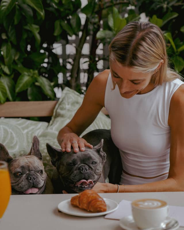 A blonde woman in a white top pats two French Bulldogs on the bench next her on, seen over a table with coffee and a pastry.