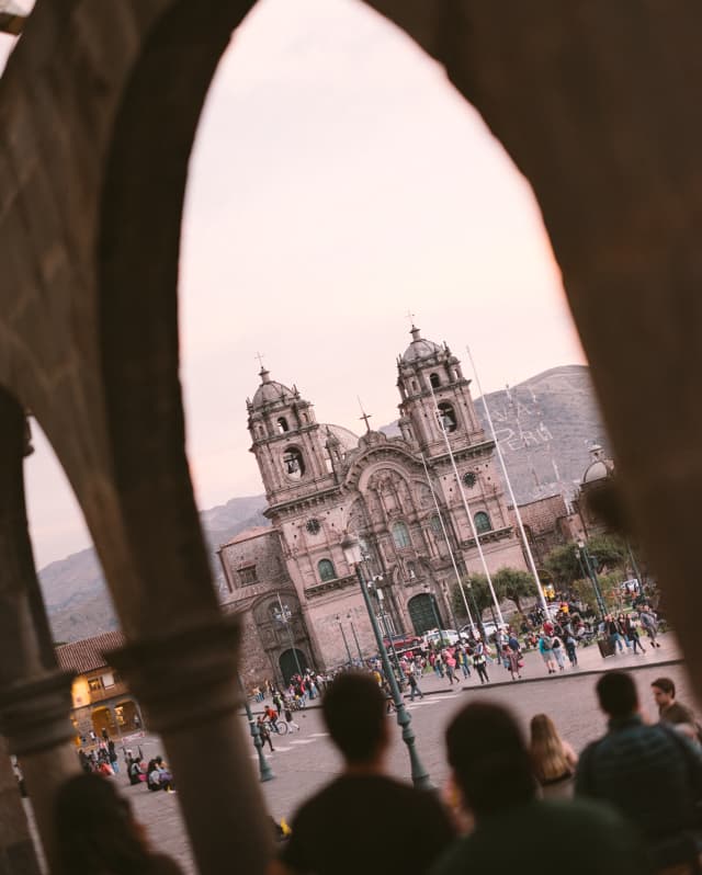 Angled low-light shot of crowds outside Iglesia de la Compañia de Jesús in Cusco's Plaza Mayor, seen through arcade arches.