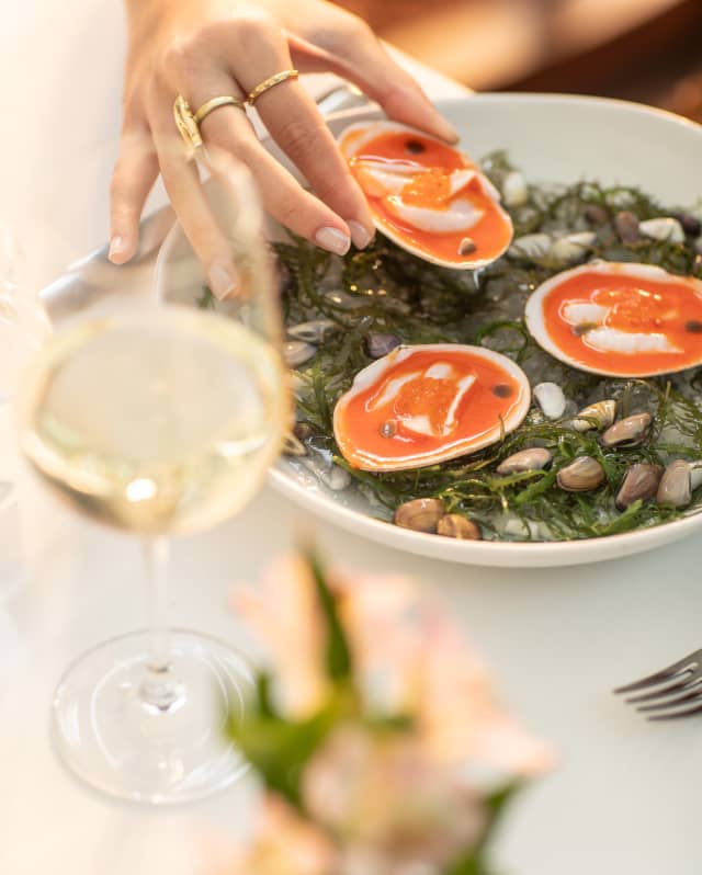 A diner's hand selects a dressed, open clam from an appetizer dish of three clams on a seaweed bed, by a glass of white wine.