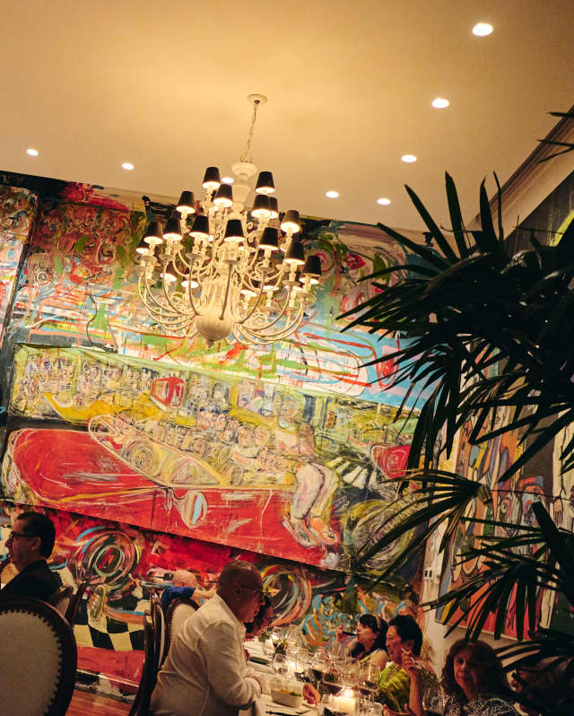 Angled image of two tables of guests as they dine beneath a chandelier and the large vivid murals of artist Mateo Liébana.