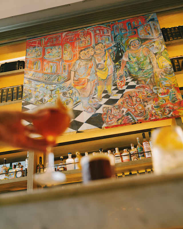 Low-angle view of a bright, busy Mateo Liébana artwork above Tragaluz bar, seen past a hand holding a drink in soft-focus.