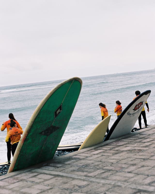 Three different-sized surfboards rest against the walkway as surfers in orange tops check out the waves, seen from behind.