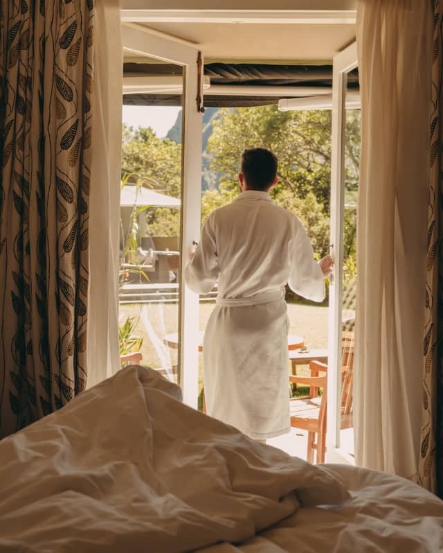 Seen across an unmade bed, a guest in a white bath robe opens the doors to his private terrace to greet the morning views.