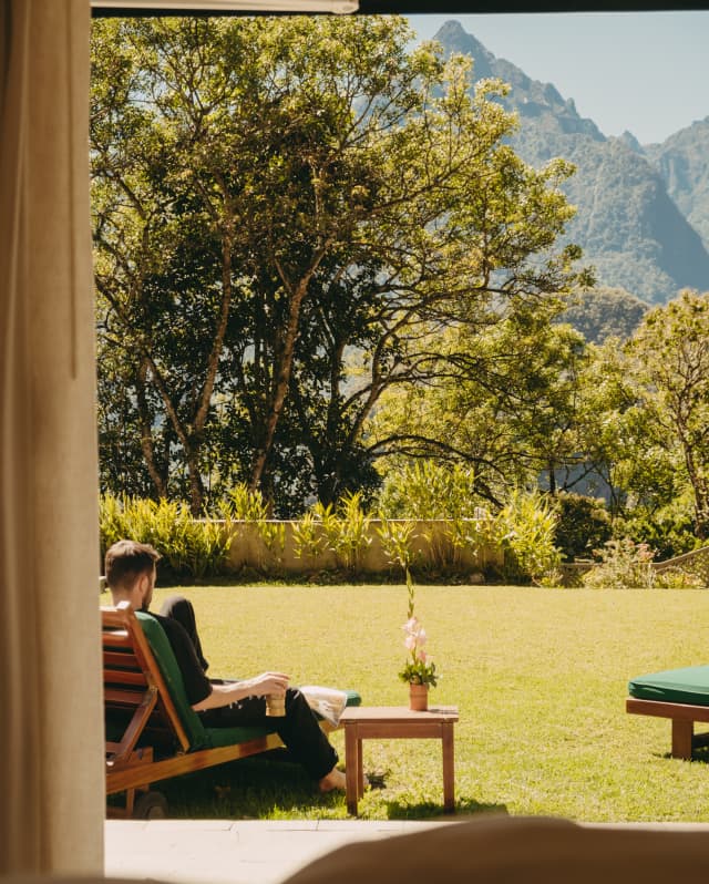 A man sits holding a drink on a sun lounger on the lawn outside his room, facing the stunning mountains, seen from behind.