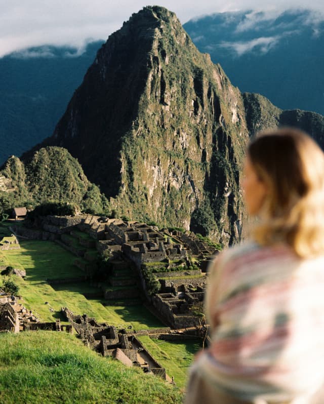In soft-focus foreground and woman sits, gazing out over Machu Picchu, with the imposing, sun-kissed Huayna Picchu in sharp focus.