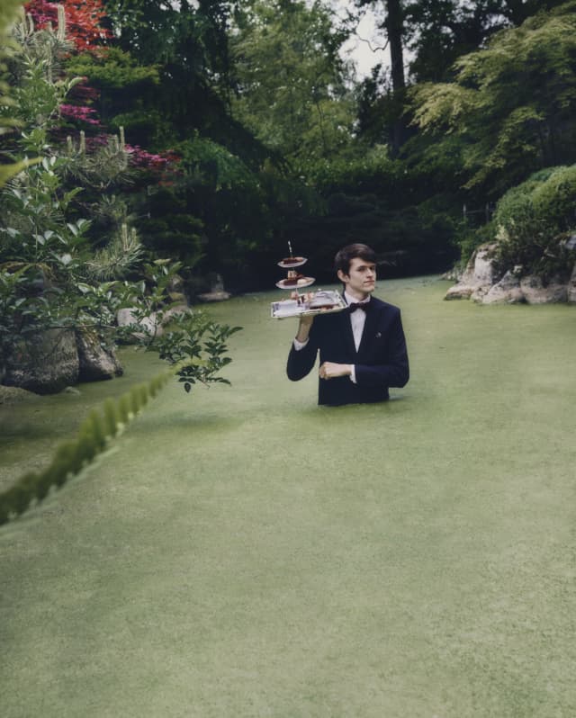 Surreal image of a waiter with a tray of afternoon tea cakes on his shoulder as he walks waist high through green pond weed.