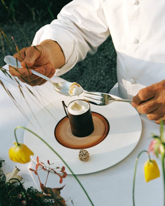 Close-up of an outdoor table as a chef holding a fork and spoon dips into the creamy froth of a café crème, seen from above.