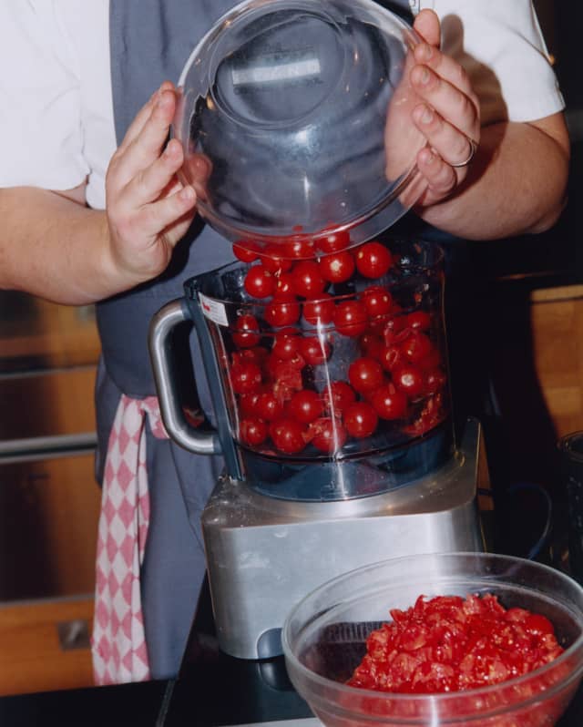 A man in a grey apron tips a glass bowl of cherry tomatoes into a food processor in a cookery class, seen from the neck down.