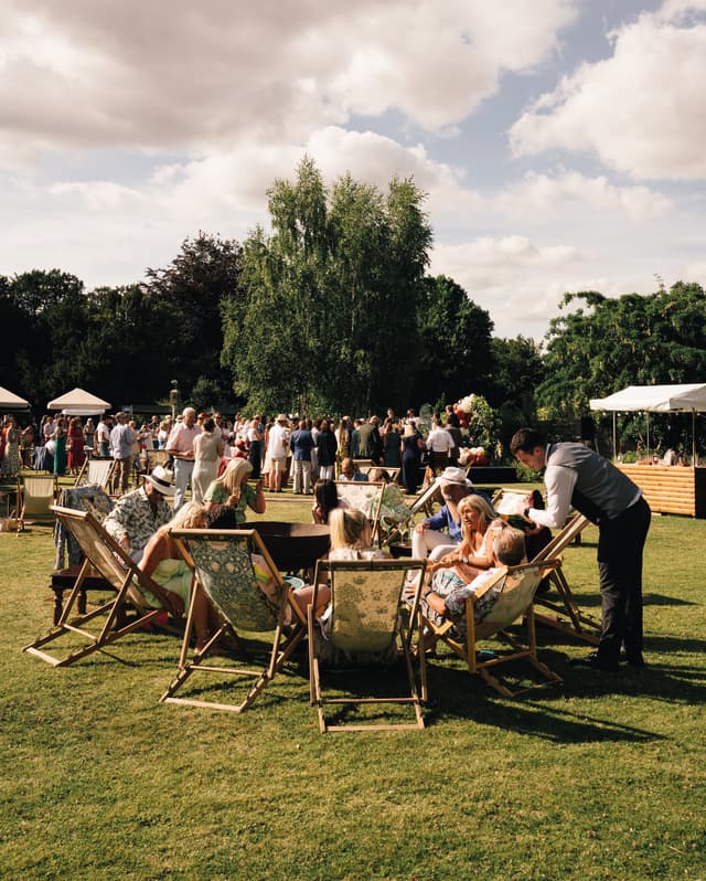 Guests sit on floral deckchairs and mill around the spacious lawn at a summer garden party with gazebos of refreshments