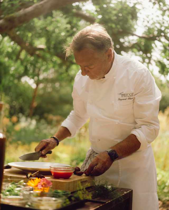Chef Raymond Blanc stands at a table of ingredients under a tree as he prepares a dish in the beautiful grounds of Le Manoir.