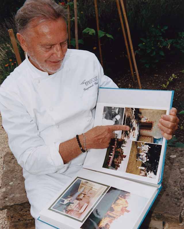 Chef Raymond Blanc, wearing chef whites, smiles as he points to old photographs in a photo album.
