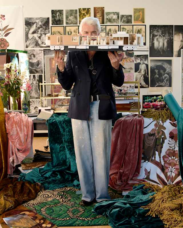 A person with grey hair stands in a creative studio, holding a model structure. The space is filled with plants, fabric, artwork, and design materials. The walls are covered in photos and sketches, creating an artistic atmosphere.