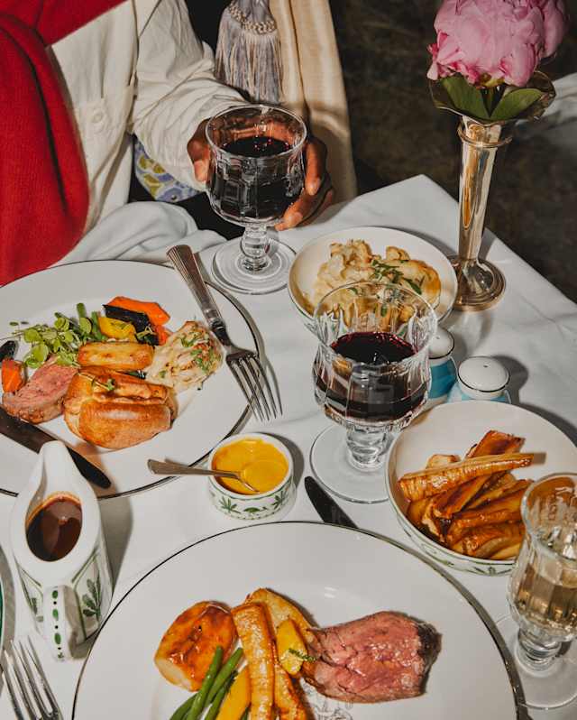 A diner is glimpsed holding a glass of red wine at a table with two plated roast beef dinners with vegetables and gravy.