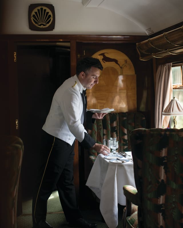 A waiter sets a table in an art deco carriage. Divine marquetry on the wall behind him shows a gazelle leaping a gold hillock
