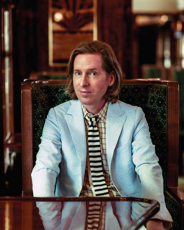 Wes Anderson smiles quizzically, dressed in a pale blue seersucker blazer, yellow and navy striped tie and yellow plaid shirt