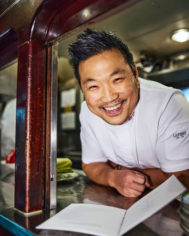 Wearing white uniform, Chef Andrew Wong smiles widely at the camera as he leans on the kitchen pass, where a menu lies open.