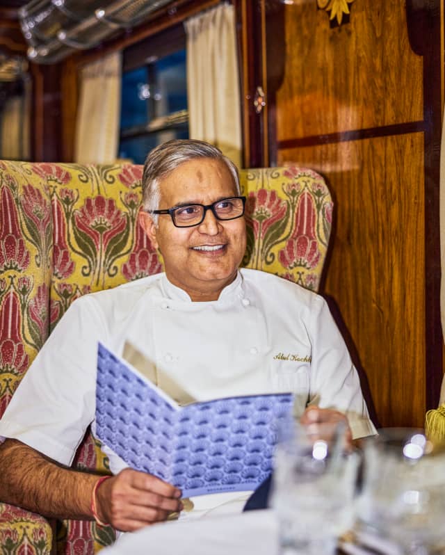 Chef Atul Kochhar, wearing black-rimmed glasses and white uniform, holds a purple patterned menu at a Gwen car dining table.