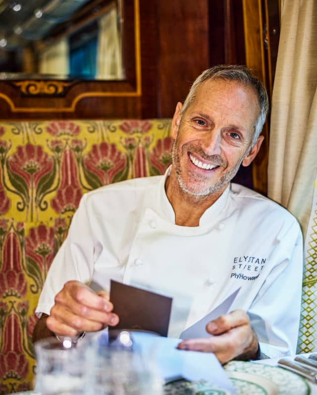 Chef Phil Howard smiles to camera, wearing whites as he sits at a table laid for dining in the Gwen carriage, holding a menu.