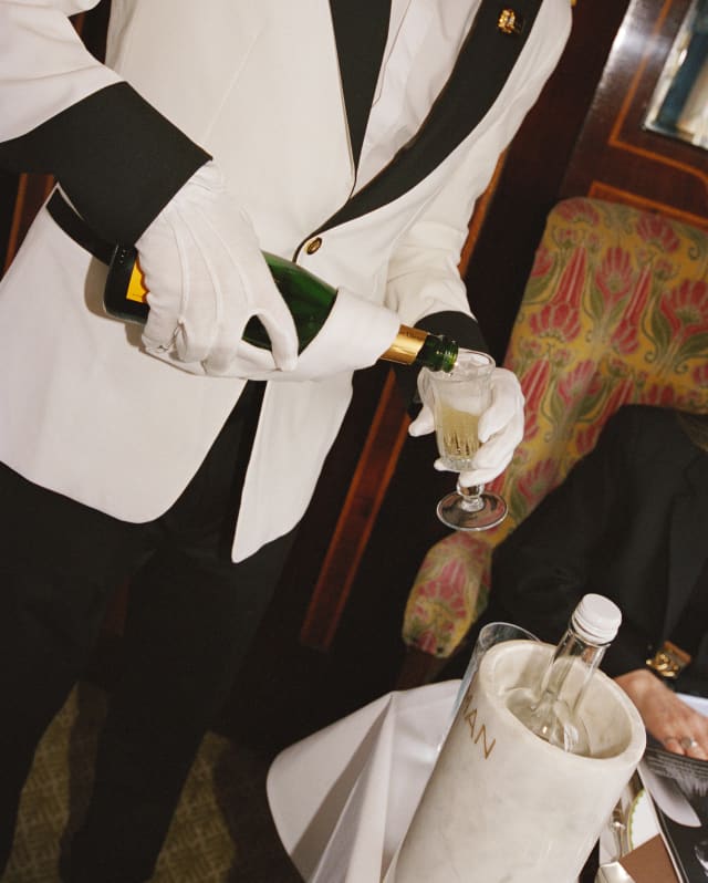 A waiter in a black and white tux pours Champagne from a bottle into a tulip glass, in an angled image taken in the Gwen car.