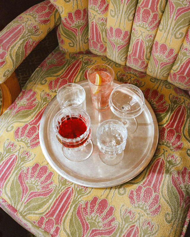 Looking down on a tray with five glasses, two with red drink, on a chair upholstered in the Gwen car's pink and gold design.