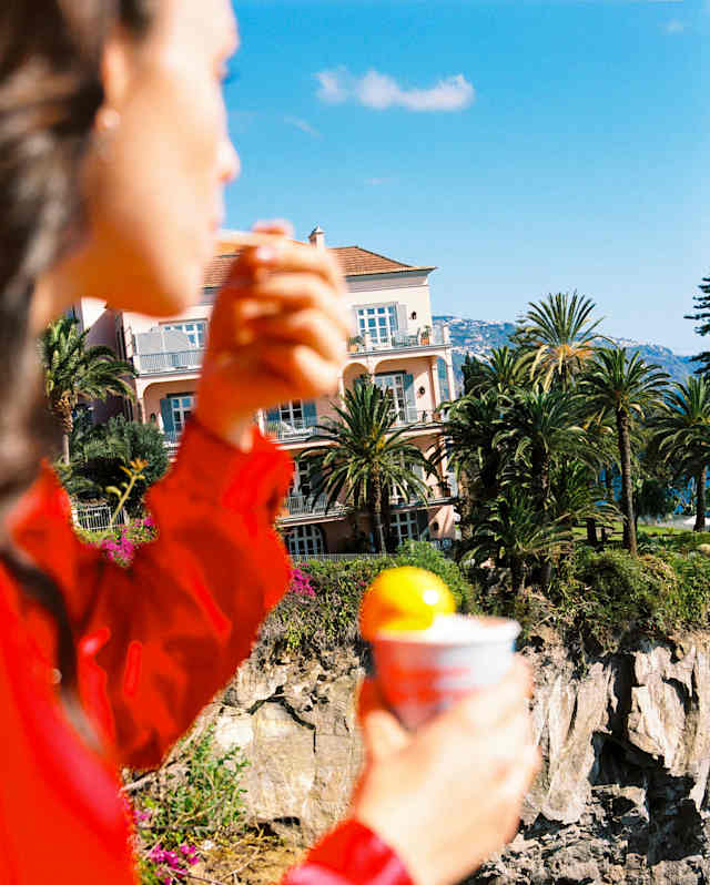 In soft-focus foreground, a woman in a red shirt enjoys a pot of orange-hued gelato, with Reid's Palace in crisp light behind.