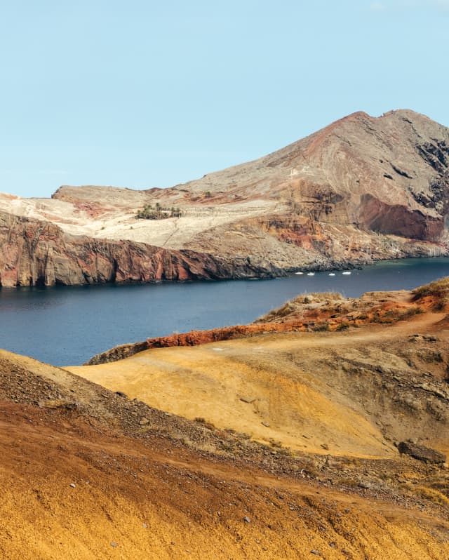 View over a rocky ridge with gold grasses to a deep blue inlet where four small boats are anchored by Ponta de São Lourenço.