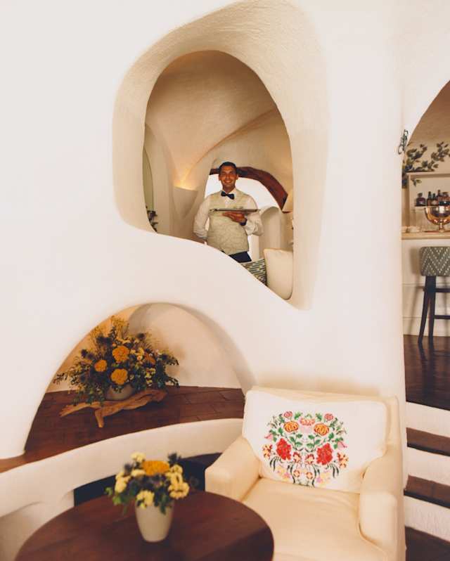 A smiling waiter, holding a tray, is glimpsed in a wall arch in a view of Bar Ginepro, capturing alcoves, armchair and table.