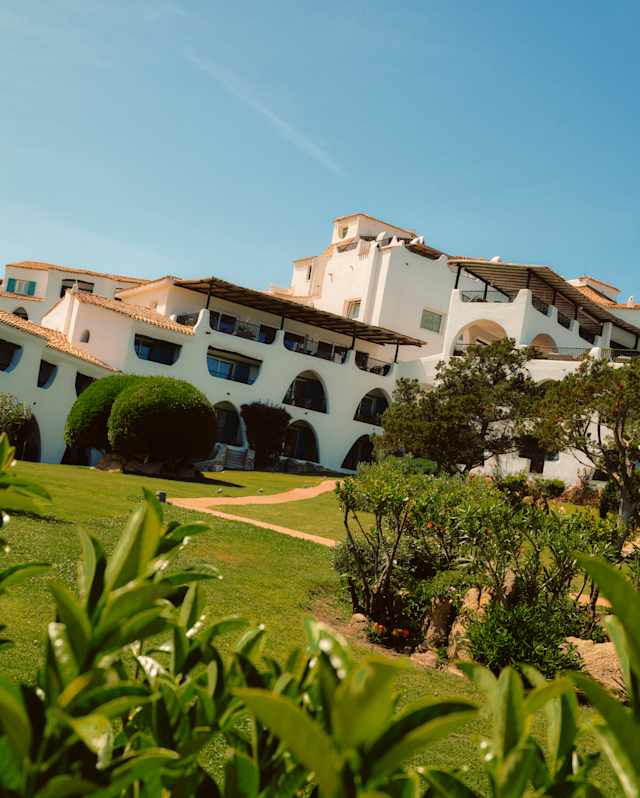 Angled image of Romazzino's white exterior with arch and rhombus windows and roof terraces, seen from lush green gardens.